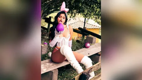 Young woman in white bunny cosplay outfit sitting on a wooden bench in a sunny outdoor park setting.