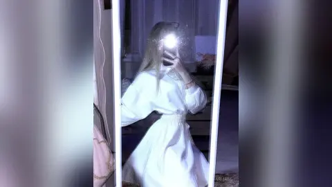 Young woman in a white long-sleeve dress taking a mirror selfie in a dimly lit room.
