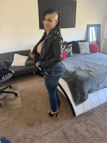 Smiling woman in a black leather jacket and blue jeans standing in a modern bedroom.