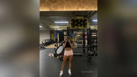 Fit woman in black crop top and pink shorts taking a selfie in a modern gym setting.