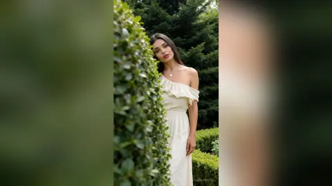 Elegant woman in a white off-the-shoulder maxi dress standing by lush green bushes in a serene garden setting.
