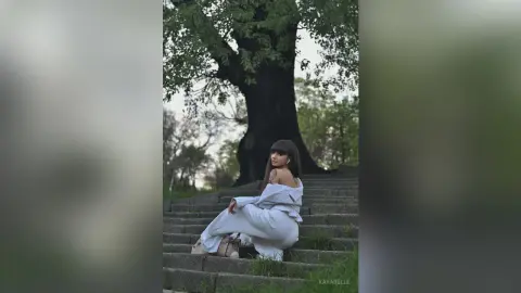 Young woman in white outfit sitting on stone stairs under a large tree in a serene outdoor setting.
