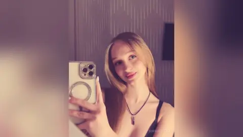 Young woman with long blonde hair taking a selfie in a black tank top with a moody, soft-lit indoor aesthetic.