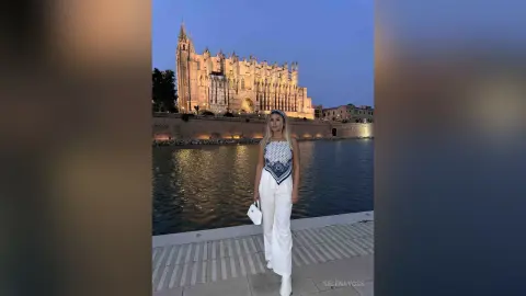 Woman in white trousers and patterned top standing by the water in front of a historic cathedral at twilight.