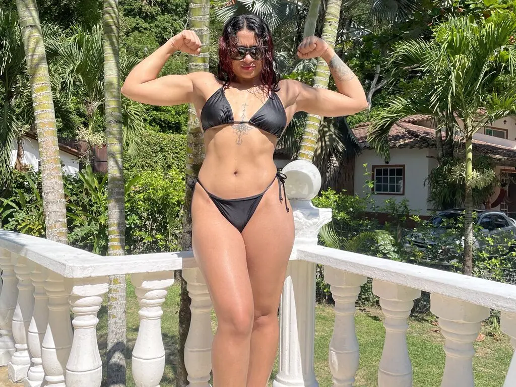 Fit woman in black bikini posing confidently on a tropical villa balcony surrounded by palm trees.