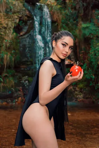 Young woman in a black sleeveless bodysuit holding a red apple near a lush forest waterfall.