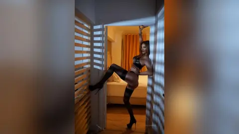 FelishaRose poses in a black bra and black stockings, leaning against a white slatted wall in a bedroom.