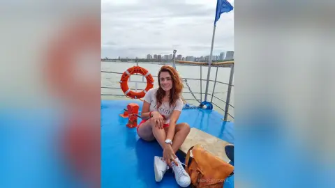 Smiling woman in a white t-shirt sitting on a blue boat deck near a lifebuoy on a cloudy day at sea.