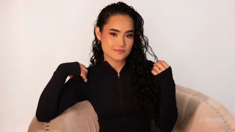 JeanRusso poses with her long, dark curly hair while wearing a black long-sleeved zip-up top, sitting in a light pink chair against a white background.