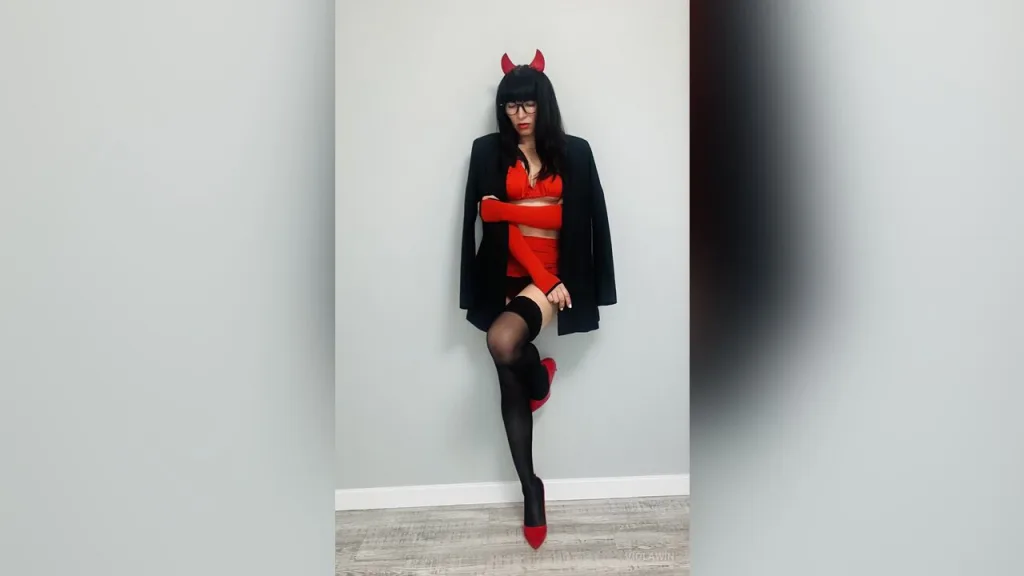 ViolaWin — ViolaWin poses in a red devil costume with horns, black glasses, and sheer black stockings against a plain wall.