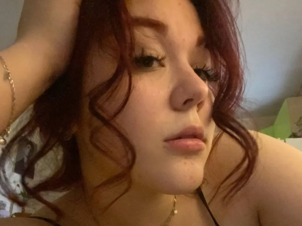 Close-up portrait of a young woman with wavy red hair and natural makeup in a soft, indoor setting.