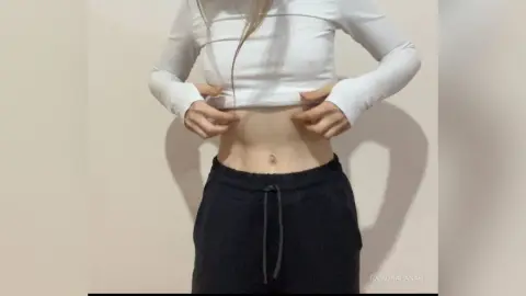 Young woman in a white long-sleeve crop top and black sweatpants posing in a minimalist indoor setting.