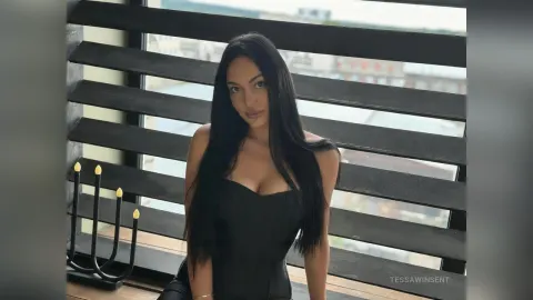 TessaWinsent — TessaWinsent poses in a black dress by a window with dark wooden slats.