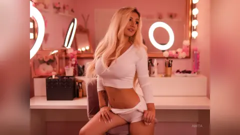 Anita poses in a white long-sleeve crop top and matching white panties in front of a lit vanity mirror.