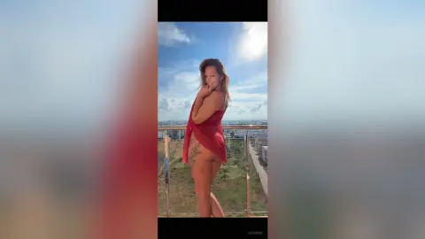 Smiling woman in a red summer dress posing on a sunny balcony overlooking a city landscape.