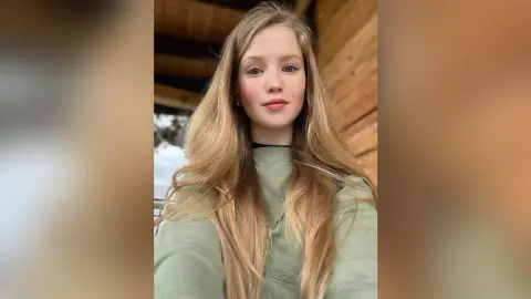 Young woman with long blonde hair wearing a sage green sweater in a cozy wooden cabin setting.