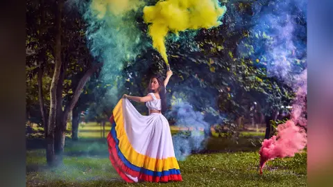 JessieLiu stands in a grassy park wearing a white crop top and a long white skirt with yellow, blue, and red stripes, surrounded by thick clouds of yellow, blue, and pink smoke.