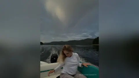Blonde woman in a white long-sleeve shirt relaxing on a boat on a calm lake under a cloudy sky.
