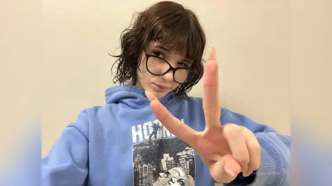 Young woman with wavy brown hair and glasses wearing a blue hoodie making a peace sign in a casual indoor setting.