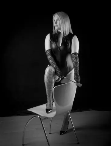 Ember_Sinn poses sitting on a white chair wearing a black lace bodysuit, fishnet stockings, and long lace gloves while holding a riding crop.