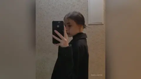 Young woman in a black coat taking a moody mirror selfie in a minimalist room.