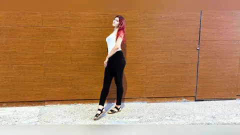 Red-haired woman in a white tank top and black leggings posing against a wooden wall background.