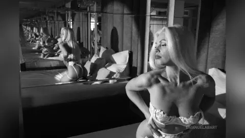 Glamorous blonde woman in white lace lingerie posing in a moody, mirrored bedroom setting.
