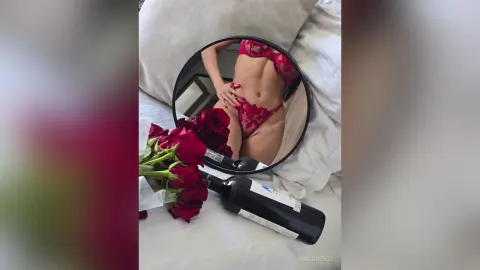 Woman in red lace lingerie posing in a mirror next to red roses on a white bed for a romantic mood.
