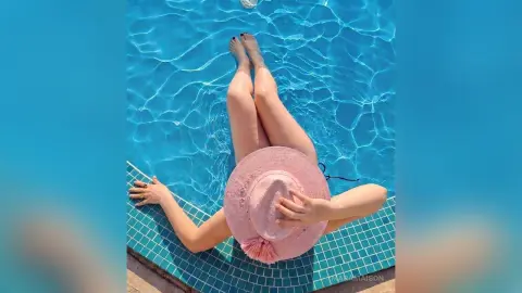 Relaxed woman in a pink sun hat lounging by a bright blue swimming pool on a sunny summer day.