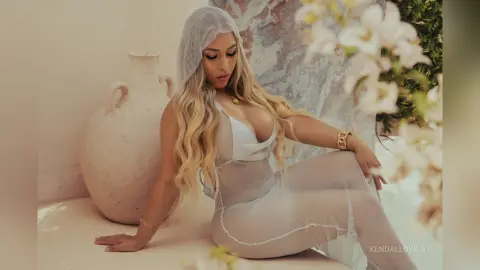 Blonde woman in a sheer white mesh outfit sitting by a large clay vase in a bright, dreamy setting.