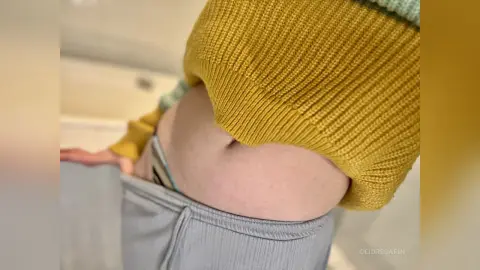 Woman in a yellow knit sweater and grey lounge pants relaxing in a bright, cozy indoor setting.