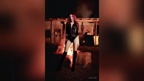 Red-haired woman in a black leather bodysuit posing in a moody, dark outdoor setting with warm firelight.