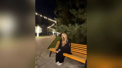 KrystalLaaz sits on a park bench at night under string lights, wearing a black outfit and white shoes.