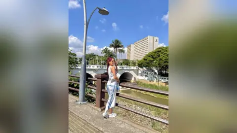 MilaGreco stands on a bridge overlooking a river, wearing a white sleeveless top and light blue denim skirt with her red hair down.