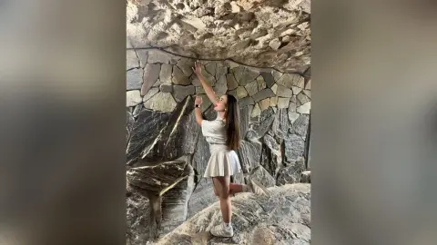 Young woman in a white summer outfit posing against a rustic stone wall in a natural cave setting.