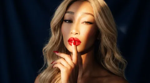 SOBLU — SOBLU poses with a finger to her red lips, featuring long blonde hair and dramatic lighting against a dark background.