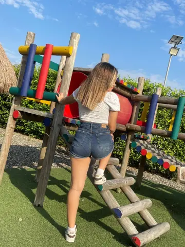 Gi_gidoll poses on a colorful wooden playground structure wearing a white crop top and tight denim shorts under a blue sky.