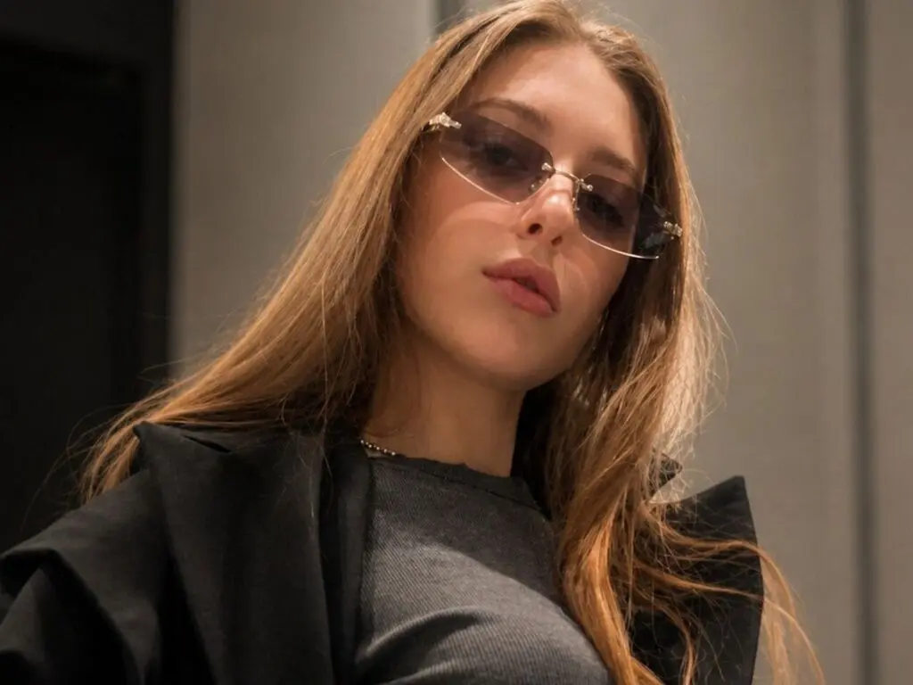 Stylish young woman in a black outfit and tinted sunglasses posing with a confident mood indoors.
