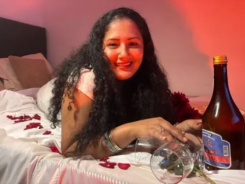 GenesisMontenegro smiles while lying on a bed covered in red rose petals next to a bottle of whisky.