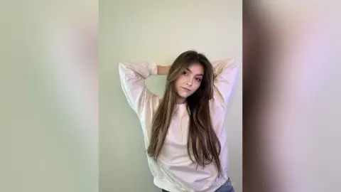 Young woman with long brown hair wearing a white sweatshirt posing against a light green wall in a relaxed mood.
