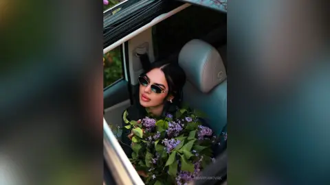 Stylish woman in sunglasses holding a large bouquet of purple lilacs while sitting in a luxury car.