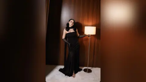 Elegant woman in a black evening gown and gloves posing in a moody, dimly lit room by a floor lamp.