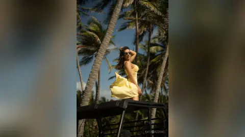 Stylish woman in a yellow summer dress posing by palm trees in a sunny tropical setting.
