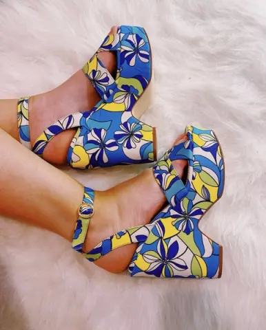 LexiLotusUS wearing blue and yellow floral pattern high-heeled sandals with ankle straps while resting her feet on a white furry rug.