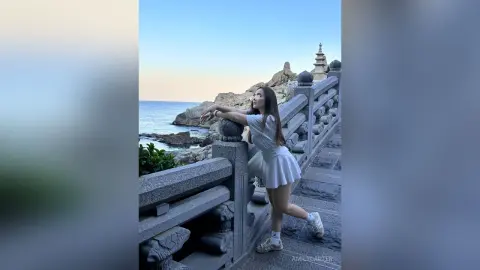Young woman in a white summer dress leaning on a stone railing by the rocky ocean coast at sunset.