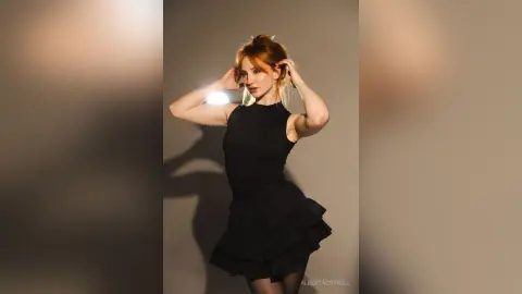 AlbertaCorrell poses in a short black dress with her hands in her red hair against a plain wall.