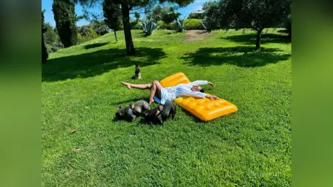 Relaxed woman in white shirt lounging on a yellow air mattress in a sunny green park with her two dogs.