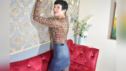 NancyTipped — NancyTipped poses in a leopard print long-sleeve top and a blue mini skirt while standing next to a red velvet sofa.