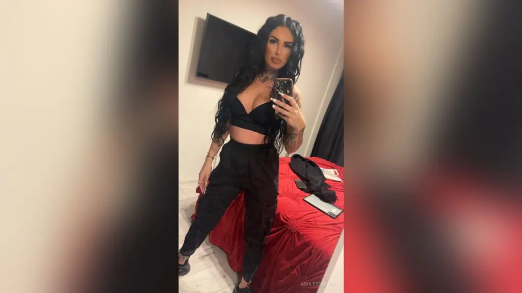 AshleighPage — Confident woman in black crop top and trousers taking a mirror selfie in a modern bedroom.