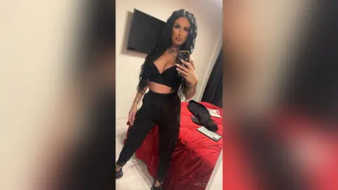 Confident woman in black crop top and trousers taking a mirror selfie in a modern bedroom.